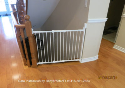 Typical installation of a white baby gate between a fixed wall and the newel post of a railing.